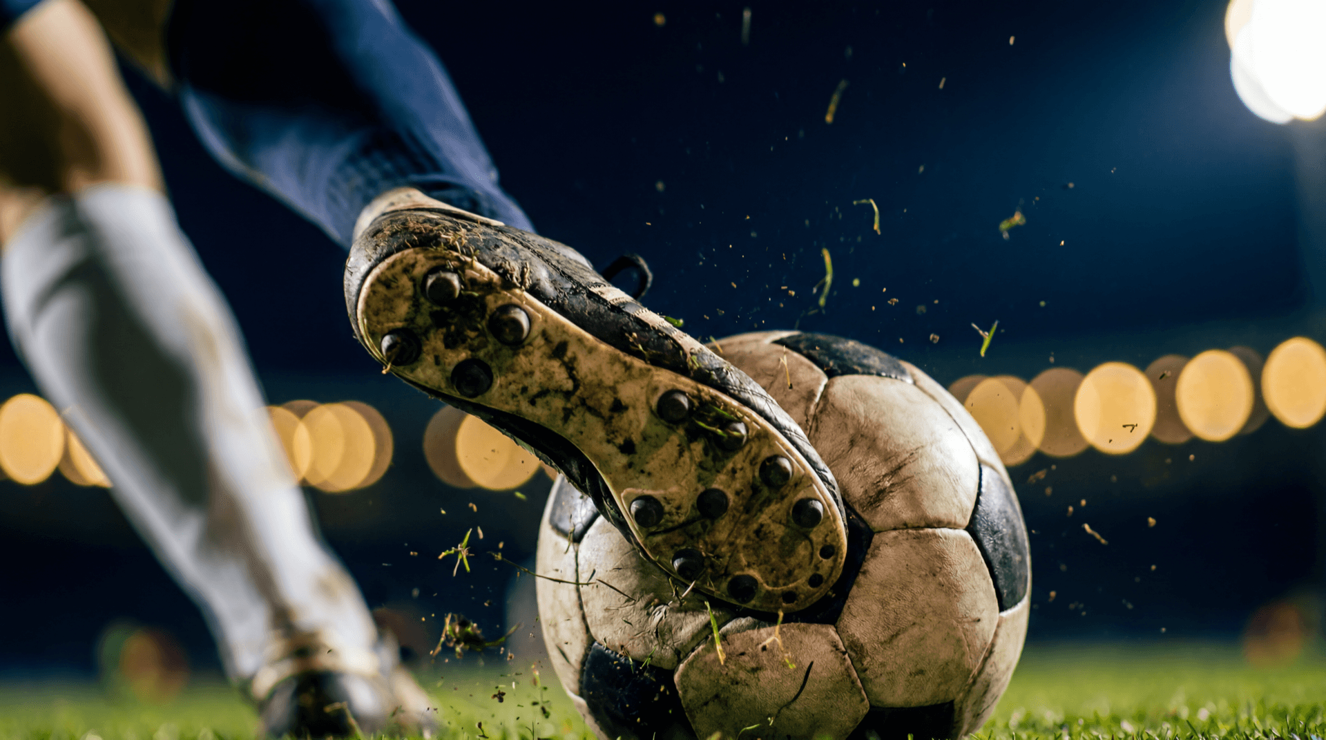 A football boot strikes a ball under stadium floodlights, grass particles flying.